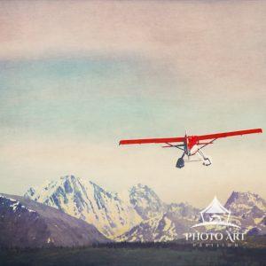 A little red float plane takes off from the water, heading toward the mountains of the Alaska Range.