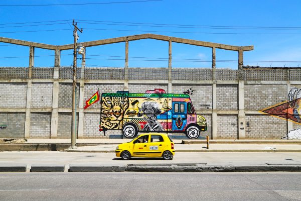 Yellow car driving past a mural of graffiti painted truck.
