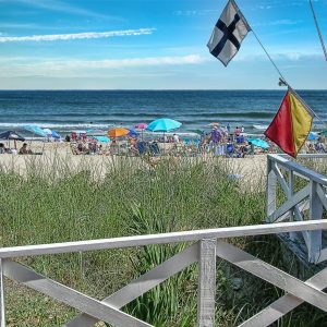 A photograph of Main Beach. In the foreground, there is a wooden fence that leads to a sandy