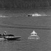 This black and white photograph depicts a water scene showing hydroplane racing. The surface of the