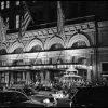 This is a black and white photograph depicting a bustling urban scene at night. The focal point is