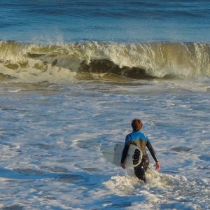 This photo depicts a dynamic ocean scene featuring a surfer, dressed in a wetsuit, standing in the