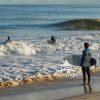 This photograph captures a serene beach scene during late afternoon. In the foreground, a surfer