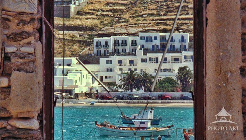  ﻿A vibrant Greek seascape is photographed through an old, rustic stone window frame. In the