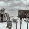 This black and white photograph depicts a coastal scene. It shows a quaint and rustic waterfront