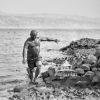 The is a black and white photograph of a man emerging from the Dead Sea covered with Dead Sea Mud.