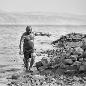 The is a black and white photograph of a man emerging from the Dead Sea covered with Dead Sea Mud.