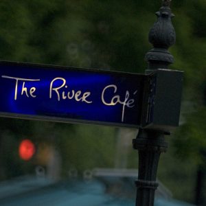 The is a photograph featuring a dark signpost with "The River Café" written in white text against