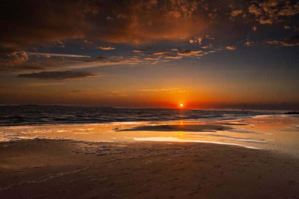 The artwork is a photograph capturing a serene beach sunset. The sun is positioned low on the
