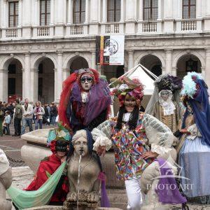 The artwork consists of a group of people dressed in elaborate, colorful costumes and masks that
