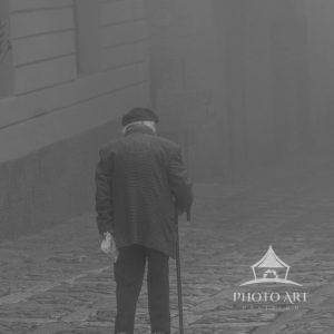 The artwork is a monochromatic photograph capturing an elderly person walking down a cobblestone