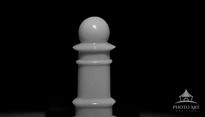 A black and white image of a solitary Pawn rises at the board's center bathed in a narrow circle of