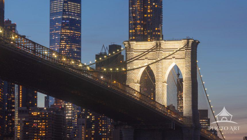 The Brooklyn Bridge glows at blue hour as it spans the East River, with One World Trade Center