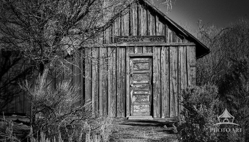 The image presents a black-and-white study of a simple wooden cabin. Vertical planks form the walls,