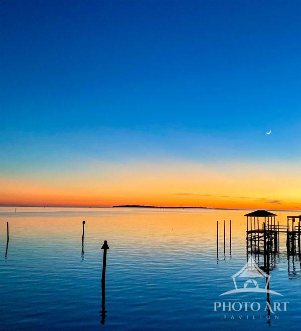 A vibrant sunset casts stunning colors across a calm body of water, with a crescent moon appearing