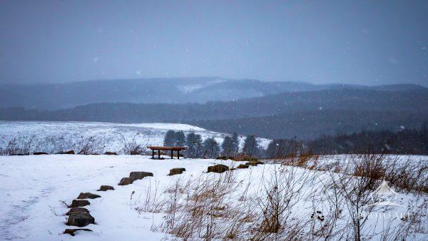 A peaceful winter scene showcasing a lone bench resting beneath falling snow, overlooking rolling