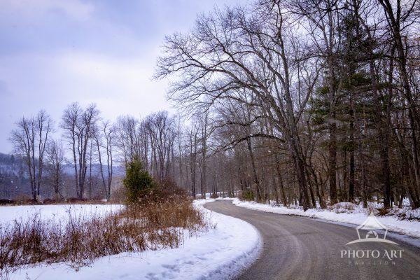This fine art photograph captures a snow-dusted country road curving through a quiet forest as