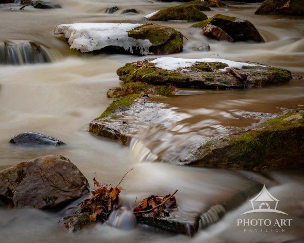 A fine art photograph capturing the gentle flow of water through a woodland stream as winter snow