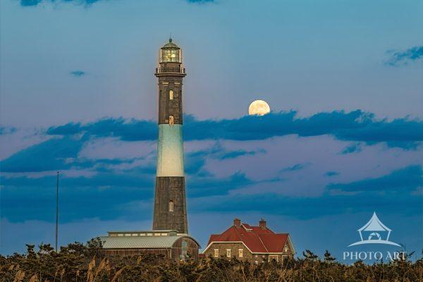 A golden full moon rises beside the historic Fire Island Lighthouse along the windswept dunes of