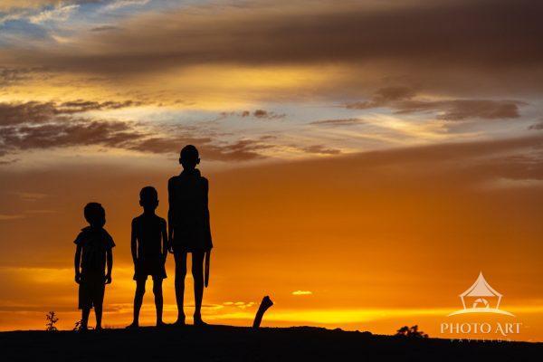 Three boys silhouetted against a sunset in Duss Village. The youngest boy looks on with admiration