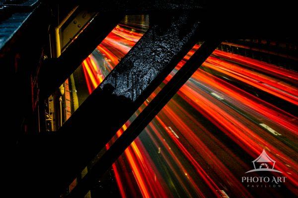 Beneath the structural beams of New York City, streaks of light carve through the darkness,