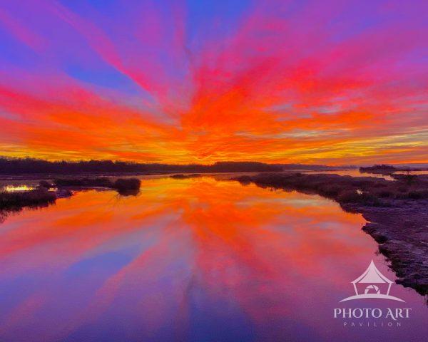 Vivid sunset paints the sky in intense orange, pink and purple hues over a calm marshland, with