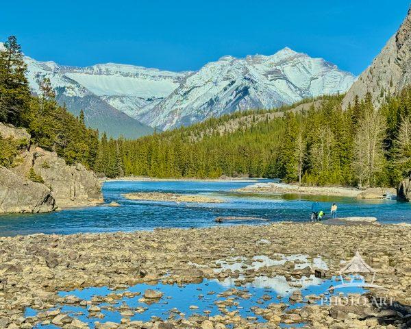 Tourists explore a rocky riverbank beneath snowcapped mountains and dense pine forest, enjoying a