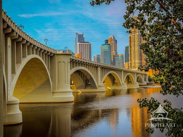 A graceful arched bridge spans the river with downtown Minneapolis skyscrapers reflected in calm