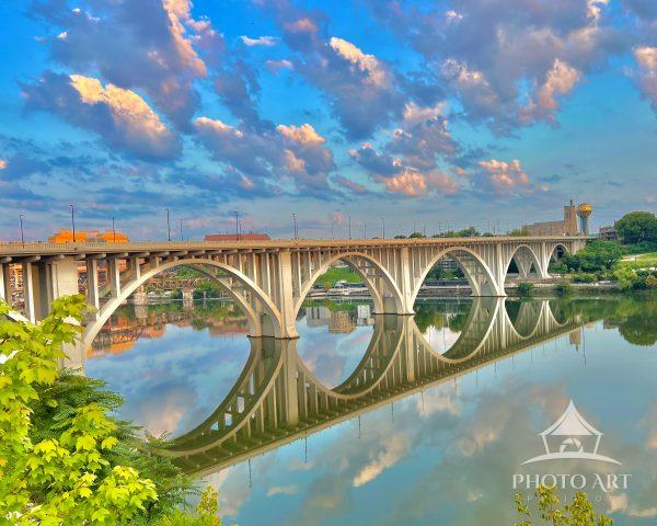 Arched concrete bridge spans a tranquil river, its arches mirrored in glassy water beneath vibrant