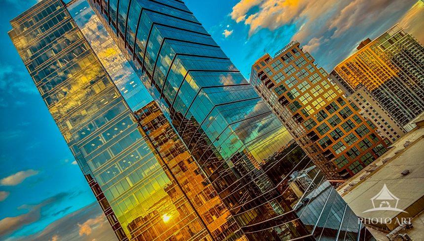 A dramatic sunset bathes a glass skyscraper and surrounding downtown buildings in golden reflection,