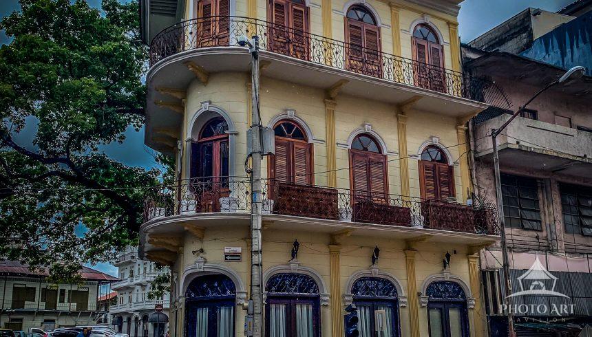 A historic three-story corner building with ornate balconies and wooden shutters overlooks a wet,