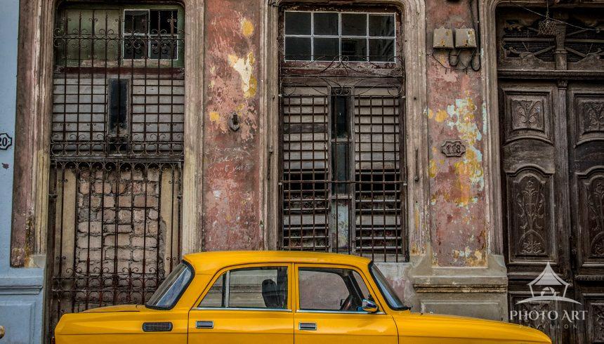 A bright vintage yellow car parked along an aged, weathered building facade with ornate doors and