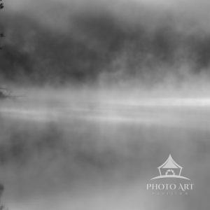 A blanket of fog drifts across still waters in this moody black-and-white scene. Dark silhouettes of