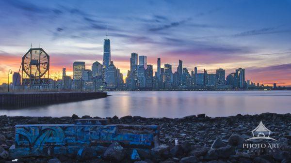 A wide landscape shot captured from Jersey City by the Colgate Clock, featuring a low-angle rocky