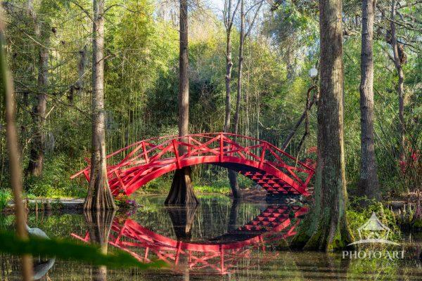 The artwork features a vibrant red arched wooden bridge spanning across a small body of water. The