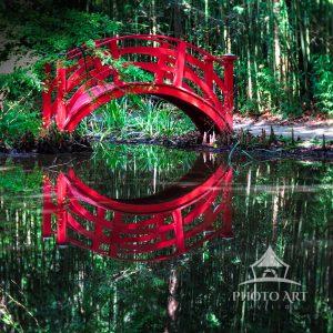 The artwork features a vibrant red arched wooden bridge spanning over a tranquil body of water in a