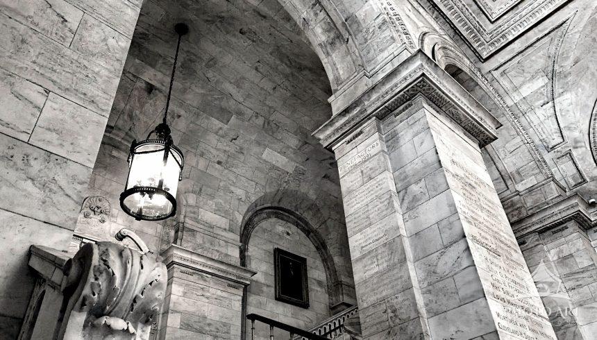 A low-angle monochrome photograph showcasing the monumental marble staircase of Astor Hall in the
