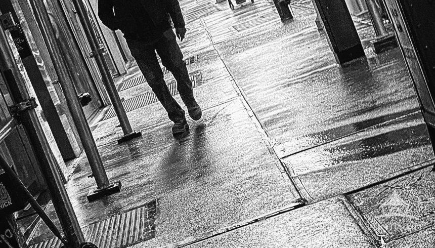 A dark, hooded figure strides down a rainy sidewalk in midtown Manhattan. Photographed with the
